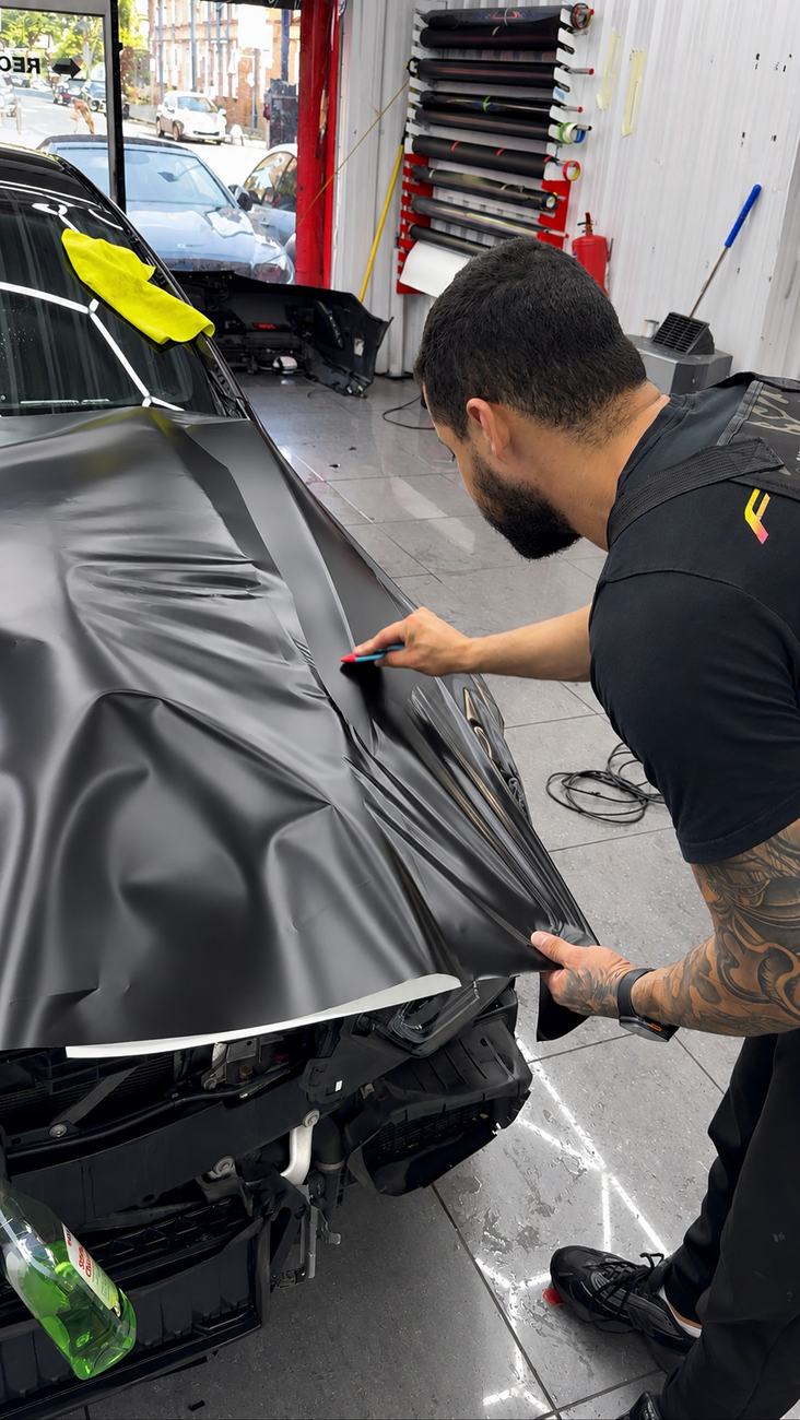 Polimax wrap technician trimming satin black vinyl on a bonnet, front bumper removed